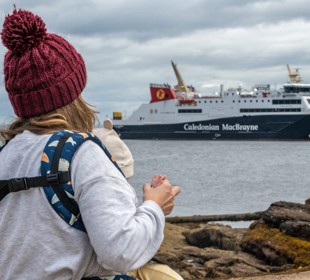 A person holding a child looks toward a Caledonian MacBrayne ferry docked near the shore on a cloudy day.