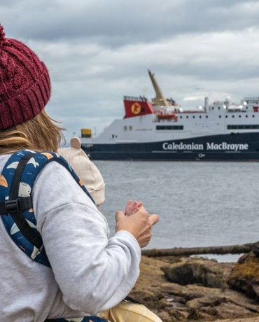 A person holding a child looks toward a Caledonian MacBrayne ferry docked near the shore on a cloudy day.
