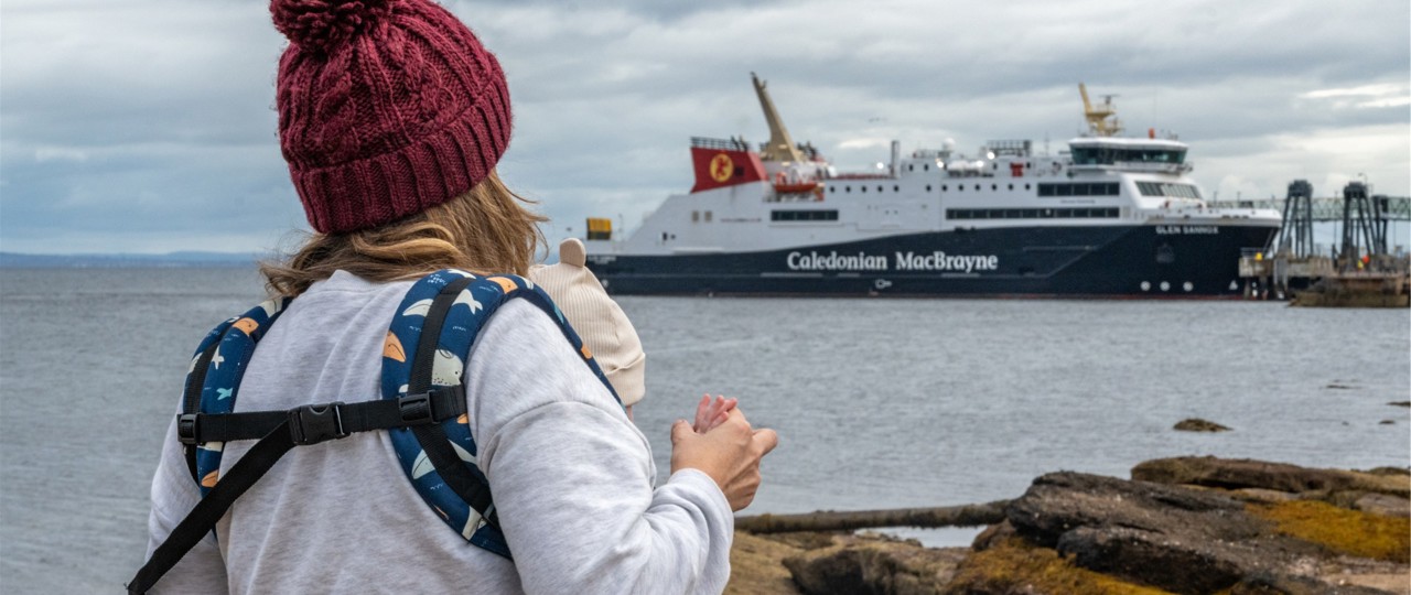 A person holding a child looks toward a Caledonian MacBrayne ferry docked near the shore on a cloudy day.
