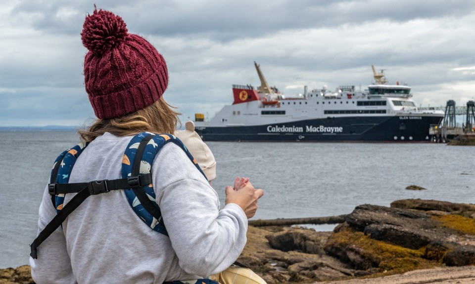 A person holding a child looks toward a Caledonian MacBrayne ferry docked near the shore on a cloudy day.