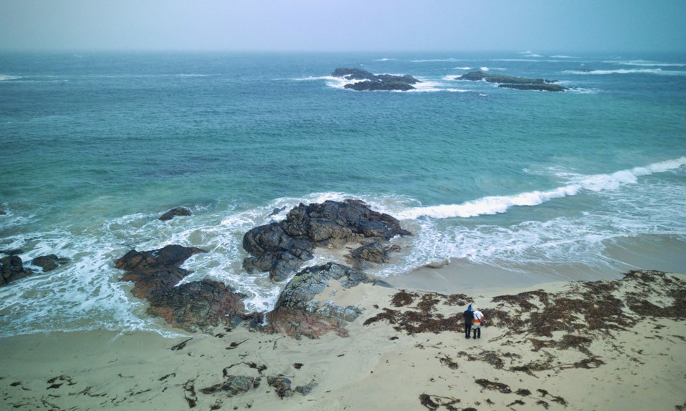 Drone overlooking Straun beach on Coll with the waves crashing against the rocks as a couple look out to sea