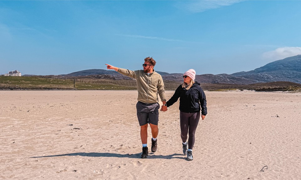 Two people walk hand-in-hand on sandy beach; one points toward horizon. Hills and house visible in background.