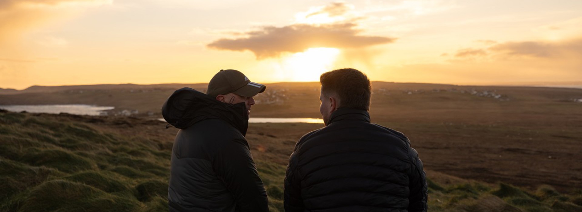 Two men in outerwear sitting on a rock on a grassy hill, facing the sunset and the island 