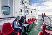 Two passengers look at a map on a ferry deck with red seats while two others stand at the railing overlooking the water.