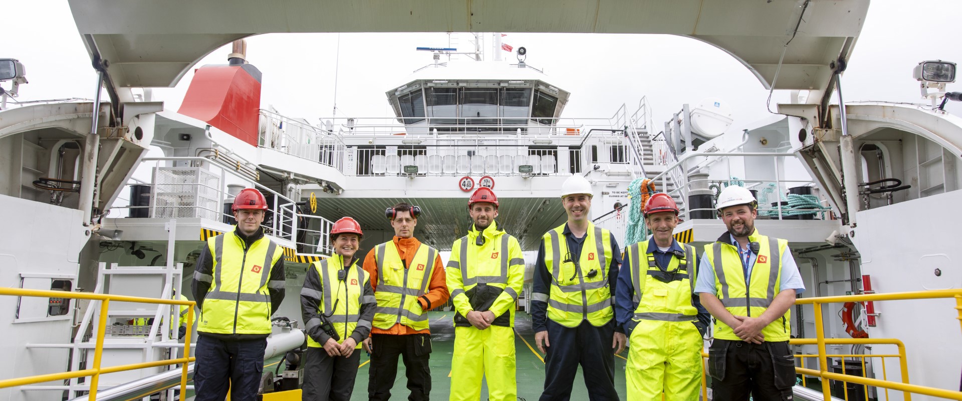 Crew members in high‑visibility gear standing on a ferry deck with the vessel’s bridge visible behind them.