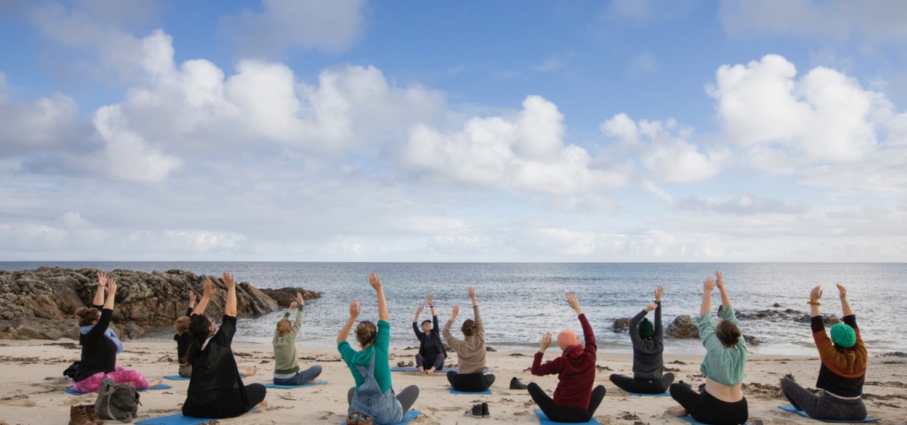 A group of people sitting on yoga mats on a sandy beach, facing the ocean. They are performing a yoga pose with their arms raised towards the sky. 
