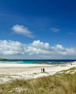 A family walking across a golden sandy beach on North Uist with dunes in the forground and blue sea and sky in the background.