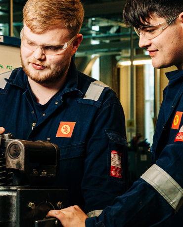 Two young men in CalMac boiler suits fixes an engine piece beside a whiteboard with technical drawings.