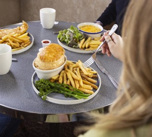 Plates of hot meals including pie, chips, greens and mugs of tea on a round table as people sit eating together.