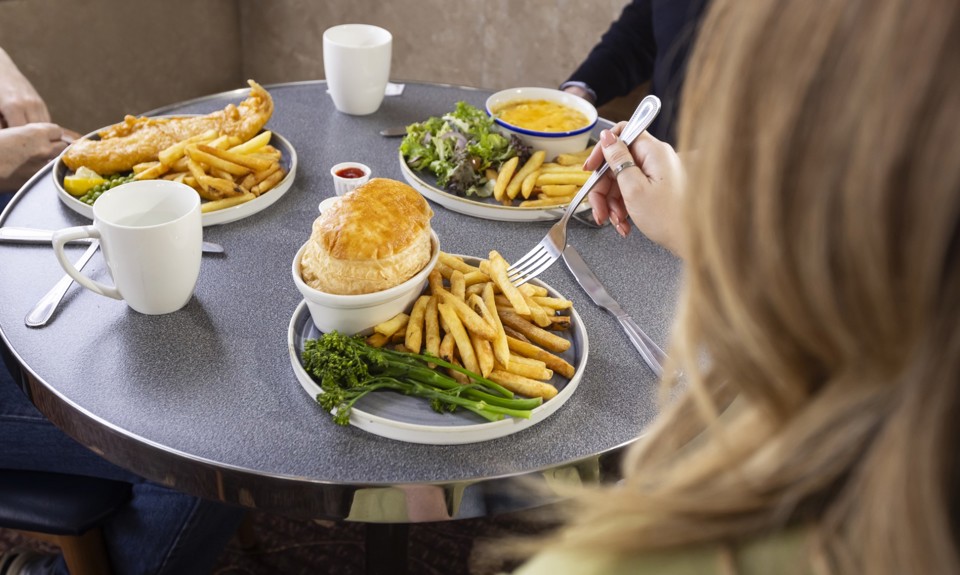 Plates of hot meals including pie, chips, greens and mugs of tea on a round table as people sit eating together.