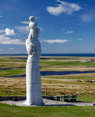 The statue called Our lady of the isles, stands proudly tall in the foreground with the flat landscape of South Uist in the background.