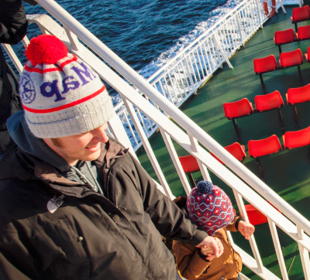 A father and child wearing winter hats walk down stairs on a ferry deck above red seats, with the sea in the background.