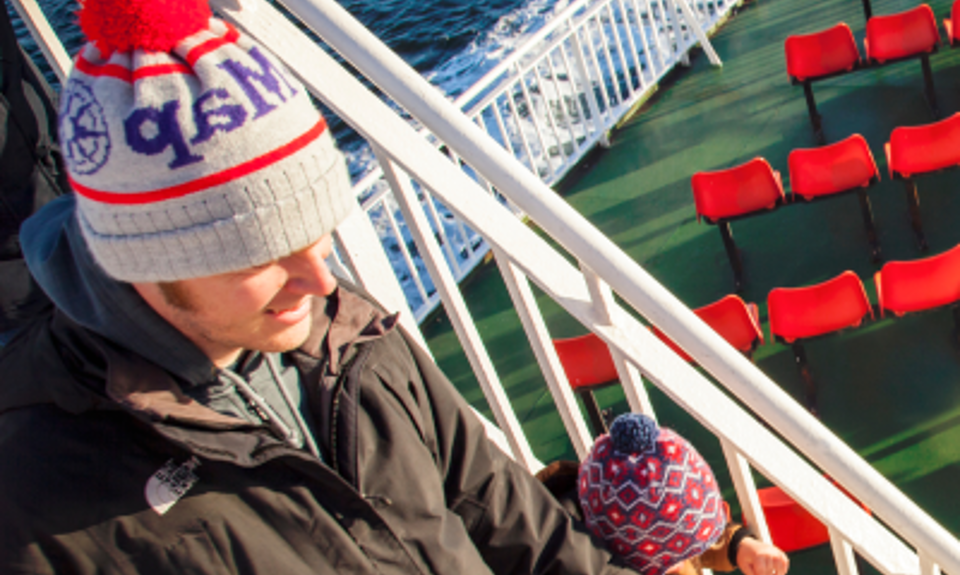 A father and child wearing winter hats walk down stairs on a ferry deck above red seats, with the sea in the background.