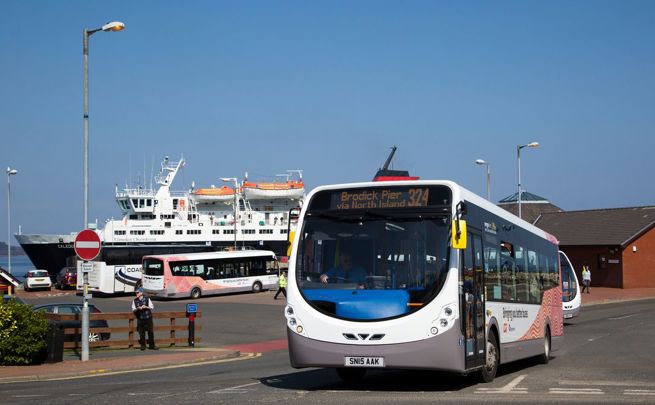 Bus 324 to Brodick Pier driving near a ferry terminal, with a large ferry and another bus visible in the background.