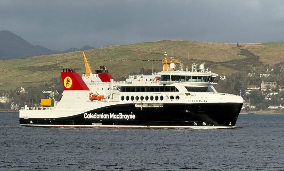 Caledonian MacBrayne ferry ‘Isle of Islay’ sailing across a calm sea with hills and houses in the background.