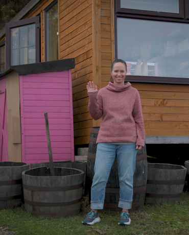 A woman standing beside a small wooden building with a pink door, surrounded by large barrel planters on a grassy area.