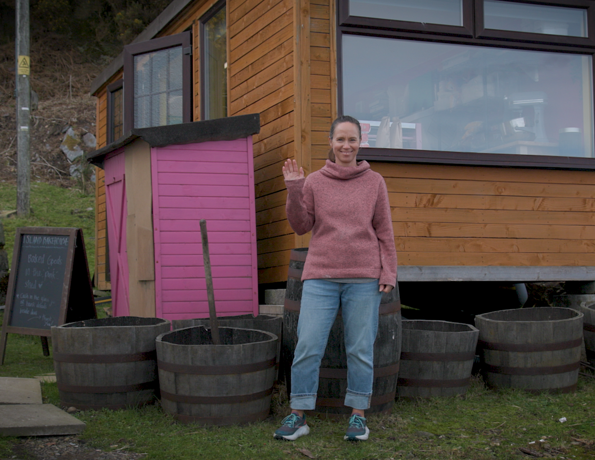 A woman standing beside a small wooden building with a pink door, surrounded by large barrel planters on a grassy area.