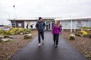 Two people walk along the path toward the Caledonian MacBrayne terminal building at Ardrossan on an overcast day.