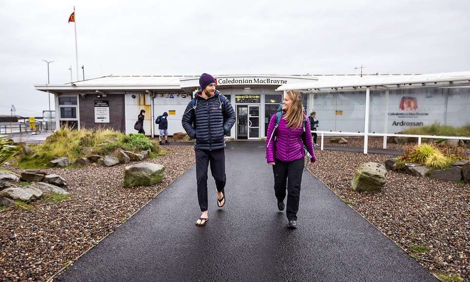 Two people walk along the path toward the Caledonian MacBrayne terminal building at Ardrossan on an overcast day.