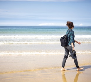 A woman walks barefoot along a sandy beach carrying shoes, with gentle waves and a blue horizon in the background.