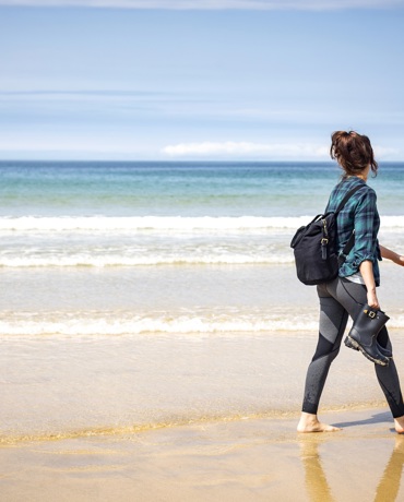 A woman walks barefoot along a sandy beach carrying shoes, with gentle waves and a blue horizon in the background.