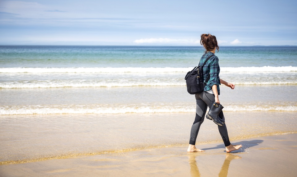 A woman walks barefoot along a sandy beach carrying shoes, with gentle waves and a blue horizon in the background.