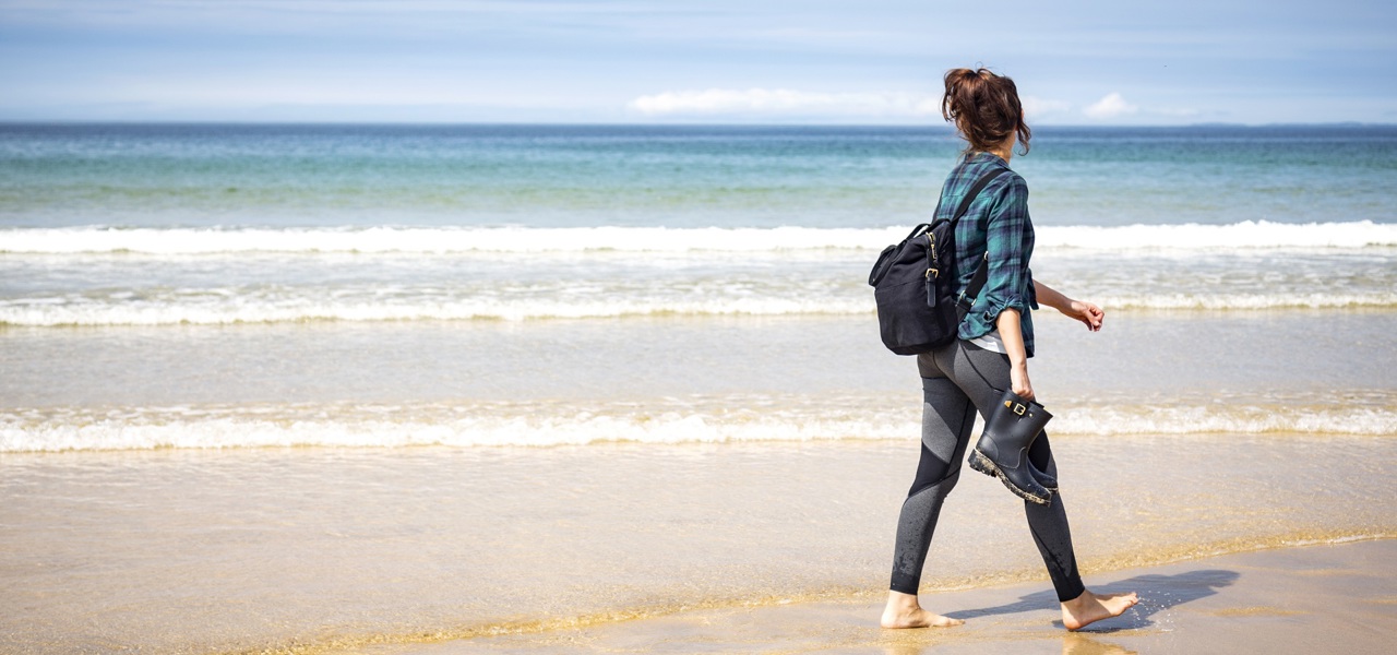 A woman walks barefoot along a sandy beach carrying shoes, with gentle waves and a blue horizon in the background.