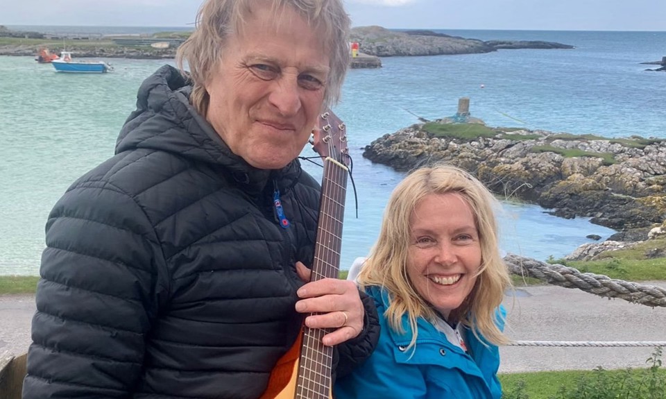 Two people by the sea, one holding a guitar, with rocky shore and boats in the background.