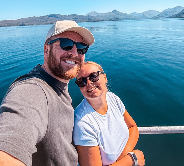 Two people on a boat with calm blue water and distant mountains in the background.