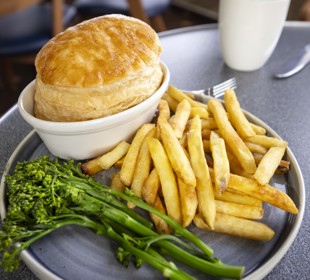 A plate of food featuring a golden pastry-topped pie, chips, and steamed greens on a table.