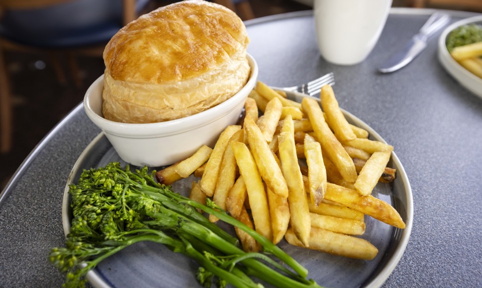 A plate of food featuring a golden pastry-topped pie, chips, and steamed greens on a table.