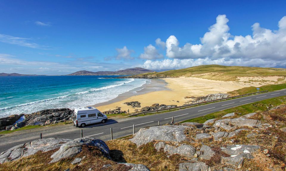 A view from a rock hill across a road to a golden beach with vibrant blue sea and sky