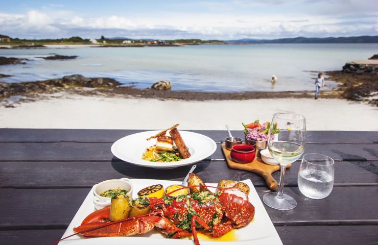 Seafood meal with lobster on a beachside table, clear skies and calm waters in the background.