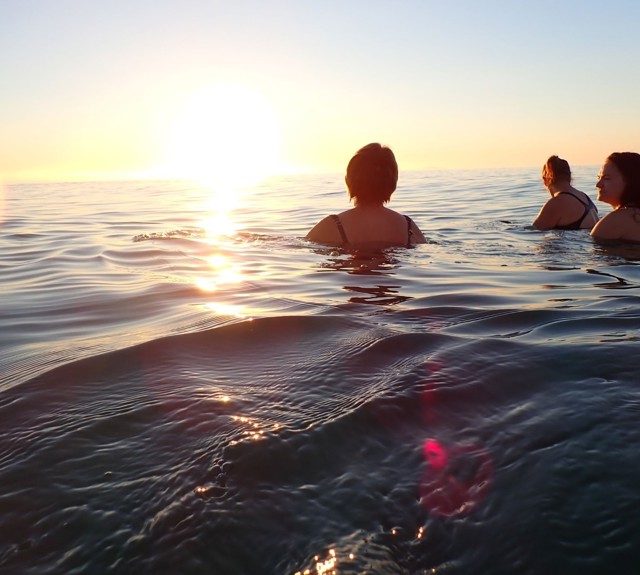 Three people swimming in calm ocean waters during sunset, with the sun low on the horizon casting a warm glow and reflections across the surface.