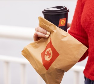woman in a red sweater holding a brown paper bag with CalMac logo and a cup of coffee in  a black cup