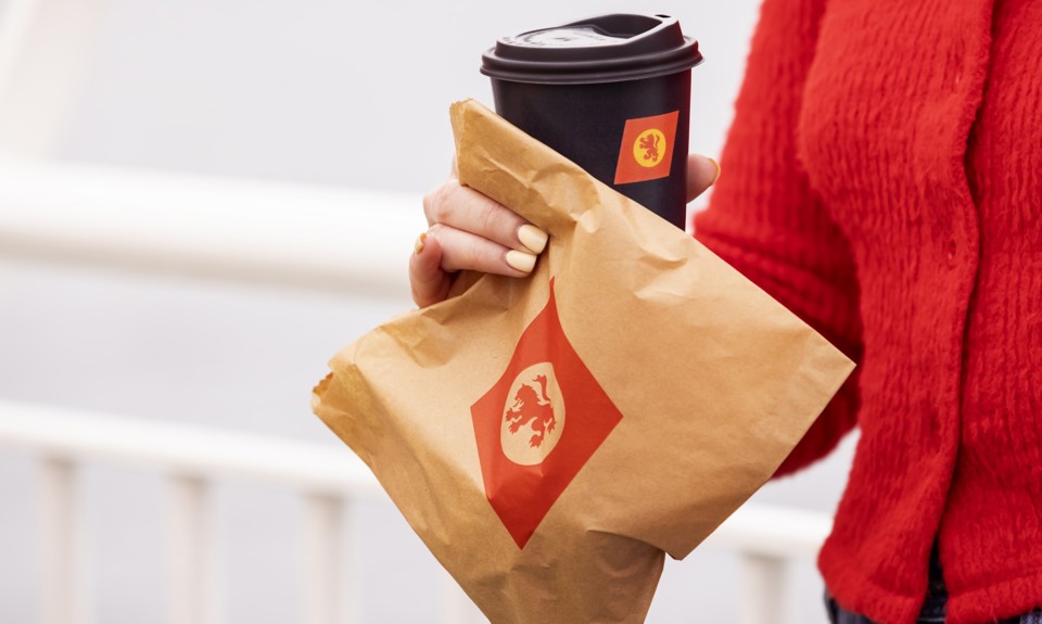woman in a red sweater holding a brown paper bag with CalMac logo and a cup of coffee in  a black cup